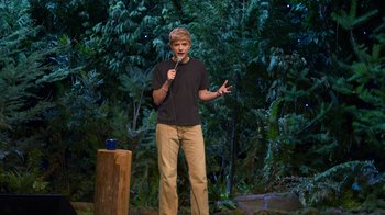 Movie still from “Mae Martin: SAP” (2023), directed by Abbi Jacobson – A man standing on a stage holding a microphone; Wide shot, High angle