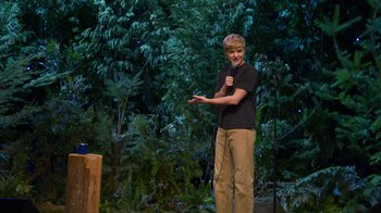 Movie still from “Mae Martin: SAP” (2023), directed by Abbi Jacobson – A young man standing on a stage holding a microphone; Wide shot, Over the shoulder angle