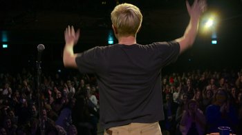 Movie still from “Mae Martin: SAP” (2023), directed by Abbi Jacobson – A man standing in front of an audience raising his hands; Wide shot, Low angle
