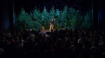 Movie still from “Mae Martin: SAP” (2023), directed by Abbi Jacobson – A man standing on a stage in front of an audience; Extreme Wide shot, High angle