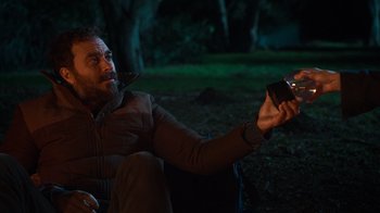 Movie still from “Mae Martin: SAP” (2023), directed by Abbi Jacobson – A man sitting in the grass holding a cell phone; Medium shot, Over the shoulder angle