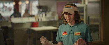 Movie still from “Maggie Moore(s)” (2023), directed by John Slattery – A man wearing a hat is standing in front of a counter; Medium shot, Over the shoulder angle