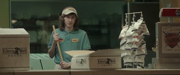 Movie still from “Maggie Moore(s)” (2023), directed by John Slattery – A person standing in front of boxes with a baseball bat; Medium shot, Low angle