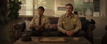 Movie still from “Maggie Moore(s)” (2023), directed by John Slattery – A couple of men sitting on top of a brown couch; Medium shot, Over the shoulder angle