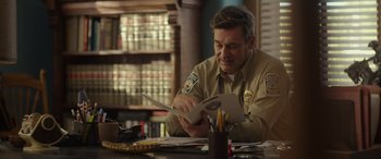 Movie still from “Maggie Moore(s)” (2023), directed by John Slattery – A police officer is reading a book at a desk; Medium shot, Over the shoulder angle