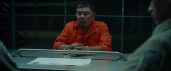 Movie still from “Maggie Moore(s)” (2023), directed by John Slattery – A man in an orange shirt sitting at a table in a jail cell; Medium shot, Over the shoulder angle