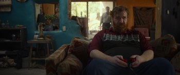Movie still from “Maggie Moore(s)” (2023), directed by John Slattery – A man sitting on top of a couch in a living room; Medium shot, Over the shoulder angle