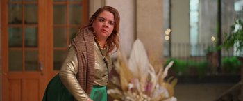 Movie still from “Making It Up” (2023), directed by Guillermo Calderón – A woman in a gold and green outfit standing in front of flowers; Medium shot, Over the shoulder angle