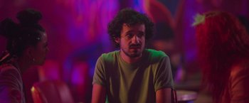 Movie still from “Making It Up” (2023), directed by Guillermo Calderón – A man with curly hair sitting in front of neon lights; Close Up shot, Over the shoulder angle
