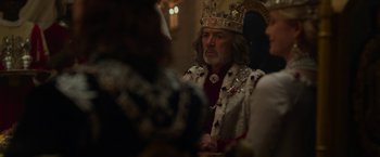 Movie still from “Maleficent: Mistress of Evil” (2019), directed by Joachim Rønning – A man wearing a crown sitting in front of other people; Medium shot, Over the shoulder angle