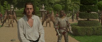 Movie still from “Maleficent: Mistress of Evil” (2019), directed by Joachim Rønning – A man in a white shirt is standing next to a man in a suit of armor; Medium shot, Over the shoulder angle