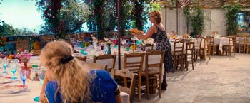 Movie still from “Mamma Mia!” (2008), directed by Phyllida Lloyd – A woman holding a tray of food in front of a table; Wide shot, Over the shoulder angle