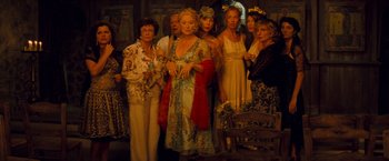 Movie still from “Mamma Mia!” (2008), directed by Phyllida Lloyd – A group of people standing around each other in front of a wooden bench; Wide shot, High angle