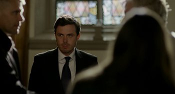 Movie still from “Manchester by the Sea” (2016), directed by Kenneth Lonergan – A man wearing a suit and tie in front of a stained glass window; Medium shot, Over the shoulder angle