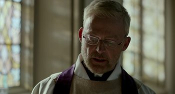 Movie still from “Manchester by the Sea” (2016), directed by Kenneth Lonergan – An older man wearing glasses and wearing a priest's outfit; Close Up shot, Low angle