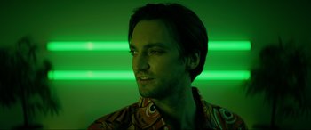 Movie still from “Margaux” (2022), directed by Steven C. Miller – A young man in a colorful shirt is in front of a neon green background; Close Up shot, Low angle