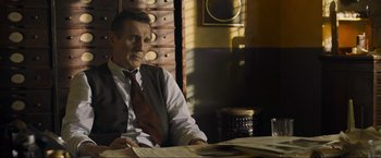 Movie still from “Marlowe” (2022), directed by Neil Jordan – A man sitting at a table in front of papers; Medium shot, Low angle