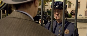 Movie still from “Marlowe” (2022), directed by Neil Jordan – A man in a police uniform is smoking a cigarette; Medium shot, Over the shoulder angle
