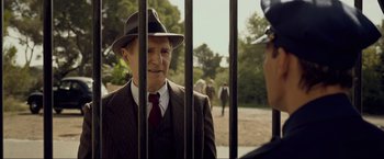 Movie still from “Marlowe” (2022), directed by Neil Jordan – A man in a suit and hat standing next to a fence; Medium shot, Over the shoulder angle