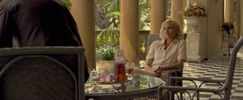 Movie still from “Marlowe” (2022), directed by Neil Jordan – A woman sitting at a glass table with a pitcher of tea; Medium shot, Over the shoulder angle