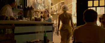 Movie still from “Marlowe” (2022), directed by Neil Jordan – A woman walking down a street past a counter; Medium shot, Over the shoulder angle