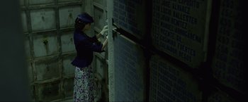 Movie still from “Marlowe” (2022), directed by Neil Jordan – A woman in a blue jacket and a hat is putting numbers on a wall; Medium shot, High angle