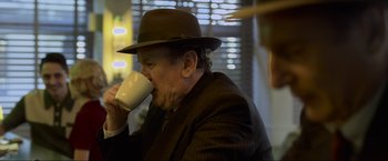Movie still from “Marlowe” (2022), directed by Neil Jordan – An older man in a hat drinking a cup of coffee; Medium shot, Over the shoulder angle