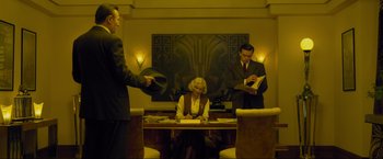 Movie still from “Marlowe” (2022), directed by Neil Jordan – A woman sitting at a desk in front of two men; Medium shot, Low angle