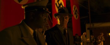 Movie still from “Marlowe” (2022), directed by Neil Jordan – A man in a suit and hat standing in front of nazi flags; Medium shot, Over the shoulder angle