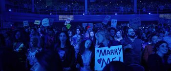 Movie still from “Marry Me” (2022), directed by Kat Coiro – A group of people in a room holding signs; Wide shot, High angle