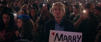 Movie still from “Marry Me” (2022), directed by Kat Coiro – A man holding up a sign that says " march " in front of a crowd; Medium shot, Over the shoulder angle