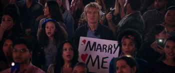Movie still from “Marry Me” (2022), directed by Kat Coiro – A man holding a sign that reads " marry me " in front of a crowd of onlookers; Medium shot, Over the shoulder angle