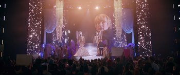 Movie still from “Marry Me” (2022), directed by Kat Coiro – A group of people standing on a stage in front of a screen; Wide shot, Over the shoulder angle