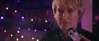 Movie still from “Marry Me” (2022), directed by Kat Coiro – A man with blonde hair is holding a microphone in his hand; Close Up shot, Low angle