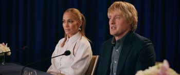 Movie still from “Marry Me” (2022), directed by Kat Coiro – A man and a woman sitting next to each other at a press conference; Medium shot, Over the shoulder angle