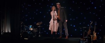 Movie still from “Marry Me” (2022), directed by Kat Coiro – A man and a woman standing next to a microphone; Wide shot, Over the shoulder angle