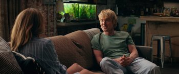 Movie still from “Marry Me” (2022), directed by Kat Coiro – A man sitting on top of a couch next to a fish tank; Medium shot, Over the shoulder angle