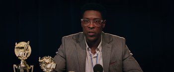 Movie still from “Marry Me” (2022), directed by Kat Coiro – A man in a gray jacket and glasses is speaking; Close Up shot, Low angle