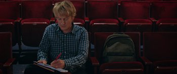 Movie still from “Marry Me” (2022), directed by Kat Coiro – A man sitting in a red chair writing on a piece of paper; Medium shot, High angle