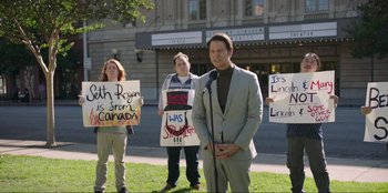 Movie still from “Maximum Truth” (2023), directed by David Stassen – A group of people standing in front of a microphone; Medium shot, Over the shoulder angle