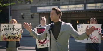 Movie still from “Maximum Truth” (2023), directed by David Stassen – A man in a gray suit is holding a microphone; Medium shot, Over the shoulder angle
