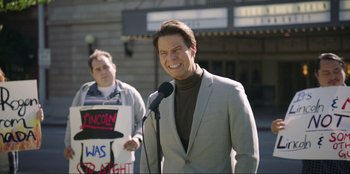 Movie still from “Maximum Truth” (2023), directed by David Stassen – A man in a gray suit is speaking into a microphone; Medium shot, Over the shoulder angle
