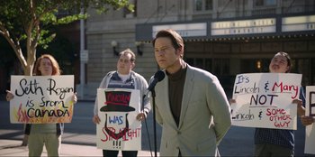 Movie still from “Maximum Truth” (2023), directed by David Stassen – A man in a gray suit is holding a microphone; Medium shot, Over the shoulder angle