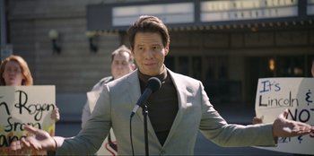 Movie still from “Maximum Truth” (2023), directed by David Stassen – A man standing in front of a microphone wearing a gray suit; Medium shot, Low angle