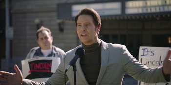 Movie still from “Maximum Truth” (2023), directed by David Stassen – A man in a gray jacket is speaking into a microphone; Medium shot, Low angle
