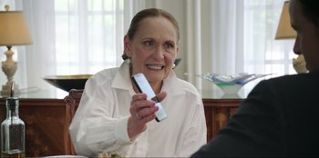 Movie still from “Maximum Truth” (2023), directed by David Stassen – An older woman holding a remote control in her hand; Close Up shot, Over the shoulder angle