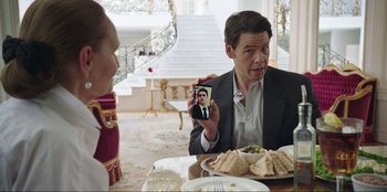 Movie still from “Maximum Truth” (2023), directed by David Stassen – A man holding up a picture of himself at a dinner table; Medium shot, Over the shoulder angle