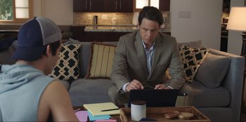 Movie still from “Maximum Truth” (2023), directed by David Stassen – A man sitting on top of a couch using a laptop; Medium shot, Over the shoulder angle