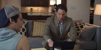 Movie still from “Maximum Truth” (2023), directed by David Stassen – A man sitting on a couch looking at a computer; Medium shot, Over the shoulder angle