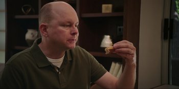 Movie still from “Maximum Truth” (2023), directed by David Stassen – A bald man holding a piece of food in his hand; Close Up shot, Over the shoulder angle